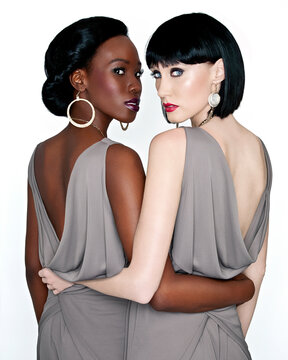 Two elegant women with stylish haircuts and makeup posing back to back in sleeveless gray dresses, with one facing forward and the other looking over her shoulder.