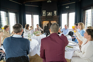 A group of people enjoying a dinner party, toasting with wine glasses around a festively set table in a room with dark wood accents and large windows.