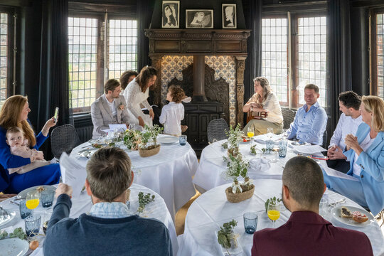 A family gathers around a dining table for a meal in an elegant room with a fireplace and classical portraits.
