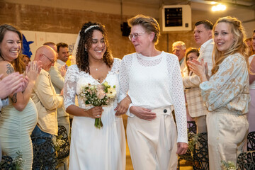 A joyous bride holding a bouquet is smiling beside an older woman at a wedding reception with guests clapping in the background.