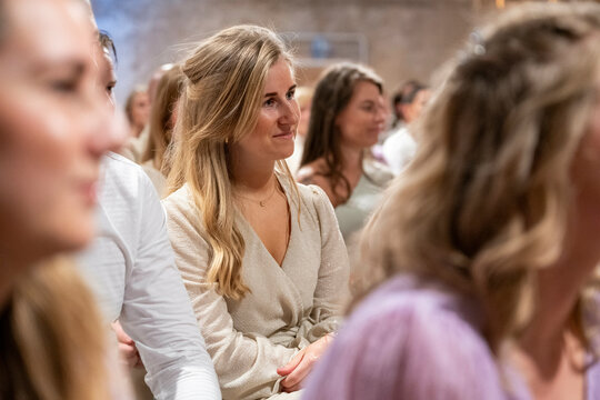 Smiling young woman in a beige cardigan sitting among a crowd of people at an indoor event.