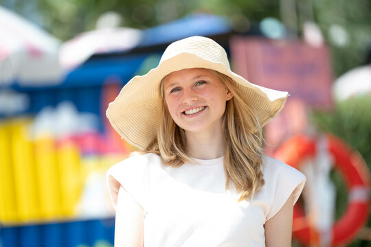 Smiling woman wearing a floppy hat in a sunny outdoor setting with blurred colorful background.