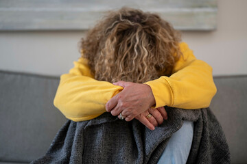 An adult with curly hair wearing a yellow sweatshirt and jeans sits hunched over on a grey couch, with their face obscured by their arms.