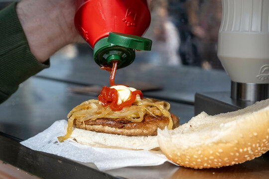 A hand squeezing ketchup onto a burger with fried onions, melted cheese, and mayonnaise on a sesame seed bun. - Powered by Adobe