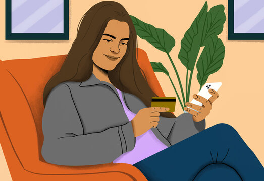 A smiling woman sits in an orange armchair, holding a credit card and a smartphone, possibly shopping online or managing finances in a cozy indoor setting.