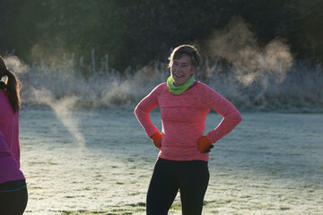A woman in a pink sports top smiles during a morning workout session on a frosty field with mist in the background and another person partially visible.
