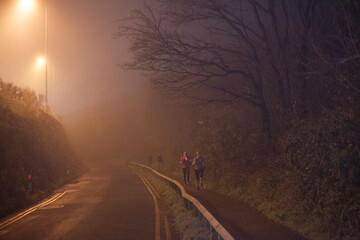 Nighttime joggers on a misty, dimly lit road bordered by trees.