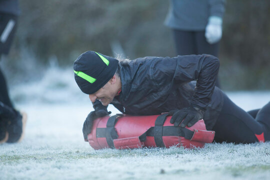 Person performing CPR on a training manikin outdoors on frosty grass with another person in the background.