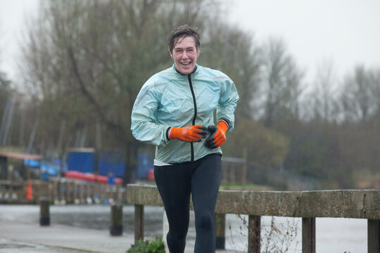 Smiling man jogging in the rain wearing a light blue jacket, black pants, and orange gloves.