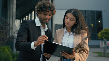 Two busy Indian business people Arabian colleagues co-workers businessman leader boss consultant and businesswoman manager executive in city downtown talking discuss document holding folder papers