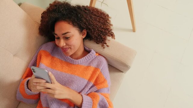 Smiling woman relaxing lying on sofa in living room browsing social media with mobile phone