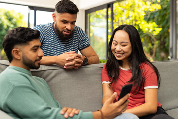 Diverse friends sitting on couch, looking at smartphone, smiling and enjoying time together