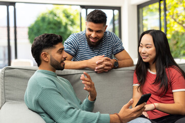 Diverse friends sitting on couch, talking and using smartphone, enjoying time together