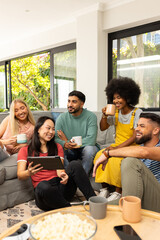 Diverse friends sitting on couch, drinking coffee, and watching tablet together, smiling