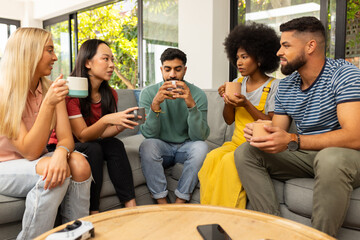 Diverse friends sitting on couch, drinking coffee, and chatting in cozy living room