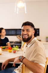 Smiling man sitting at kitchen counter with diverse friends preparing food in background, copy space