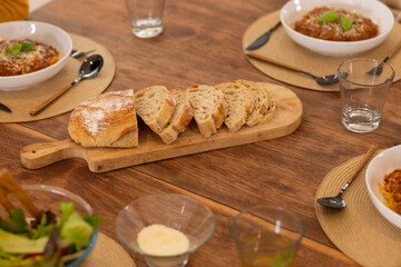 Sliced loaf of bread on a wooden table, with bowls of pasta dinner