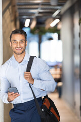 Smiling businessman holding smartphone and carrying bag in modern office hallway