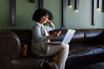 Obraz premium Using smartphone and laptop, businesswoman working remotely on leather couch