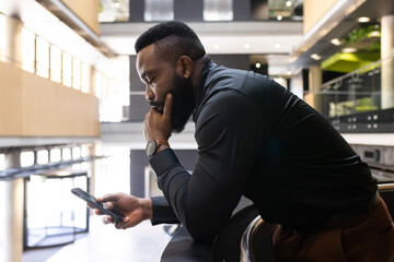 Leaning on railing, african american businessman using smartphone and thinking in modern office