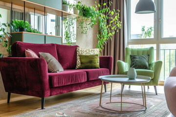 A stylish living room featuring maroon sofa and green armchair, surrounded by lush plants and natural light. cozy atmosphere invites relaxation and comfort