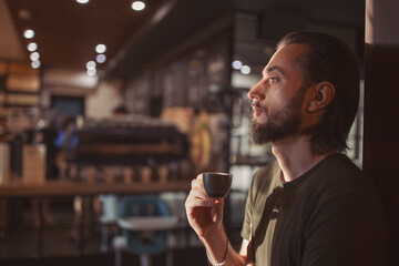 Pensive bearded hipster guy enjoying morning coffee for breakfast looking away in cafeteria, thoughtful caucasian male in casual outfit drinking beverage during break in cafe interior