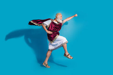 Obraz premium Full size photo of powerful mature man raise fist flying running wear ancient roman emperor tunic isolated on blue color background
