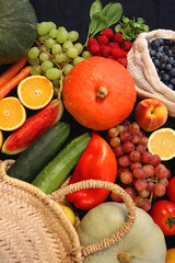Round straw bag and various seasonal fruits and vegetables on dark background. Summer and fall produce. Top view.