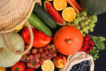 Round straw bag and various seasonal fruits and vegetables on dark background. Summer and fall produce. Top view.