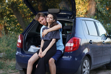 young couple sits together in the open trunk of a car parked in the forest. They are surrounded by trees and green vegetation