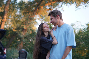 Fototapeta premium woman in a black shirt embraces a man in a blue shirt as they stand in a forest. The sun shines behind them, creating a warm, golden glow