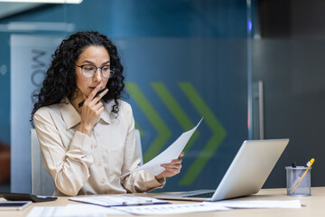 Confident businesswoman examines report while working at desk with laptop. She looks pensive,...