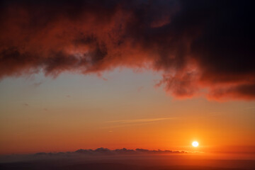 Fiery red and black clouds dominate a dramatic sunset sky, with the sun glowing on the right and a small patch of clear sky on the left, setting a striking backdrop