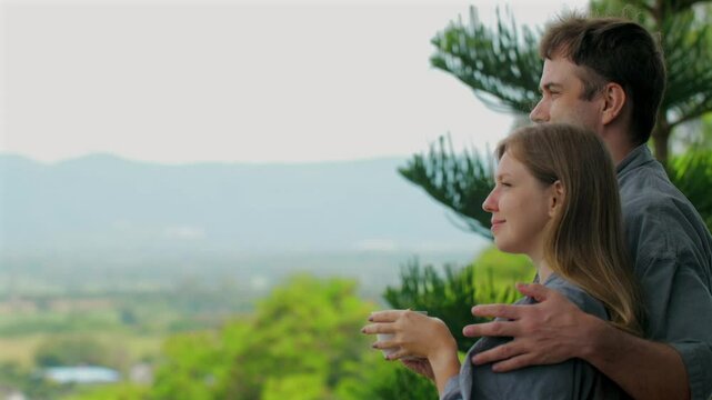 Loving couple enjoying nature view from scenic location. man and woman are casually dressed, standing together on balcony, embracing and appreciating serene landscape of mountains and trees.