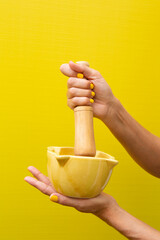 Hands holding a yellow ceramic mortar with her wooden mallet on a yellow background.
