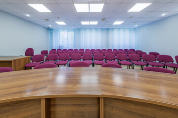empty auditorium with tiered wooden seating and a bright ceiling featuring multiple square lights. Large windows with vertical blinds are on the left side, giving the room a clean, modern atmosphere