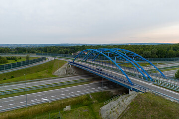 Aerial view of a modern blue arch bridge over a highway surrounded by green fields and trees, showcasing infrastructure development and nature in harmony.