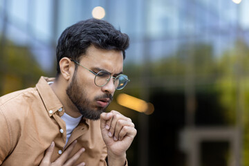 Young man coughing with hand on chest showing signs of discomfort or cold. Image captures concern,...