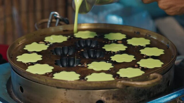 Thai street food seller pouring dough into indented pan with floral molds for khanom krok bai toei baking. Human cooking pancakes made of tapioca starch and pandas leaves with traditional method.