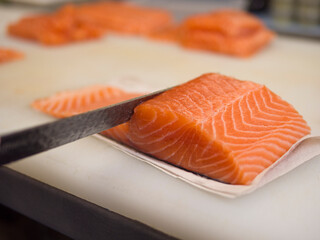 On a kitchen board, a piece of salmon is expertly sliced with a knife. The salmon's vibrant color and smooth texture shine through with every precise slice, highlighting the freshness of the salmon.