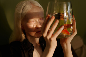 Medium closeup of young woman with albinism holding aquarium in her hands and looking at small goldfish