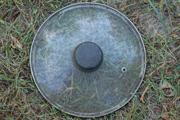 one old gray round glass dirty pot lid lies on the ground and grass on a summer street