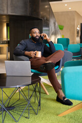 Businessman sitting in modern office, talking on phone and holding coffee mug