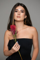 Front portrait of a beautiful young woman with makeup and attitude, wear black dress with bare shoulders, posing in studio with a red flowers.