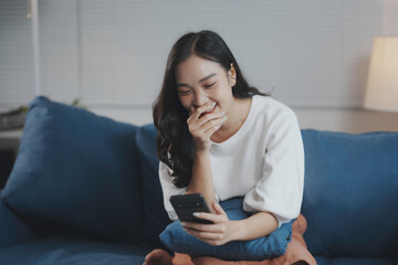 Young asian woman relaxes on her couch, laughing at something amusing on her smartphone