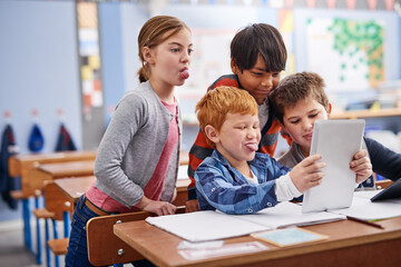 Obraz premium Funny face, selfie and tablet with student children in classroom at school together for education. Development, memory and tongue out with friends at desk in class for future, growth or scholarship