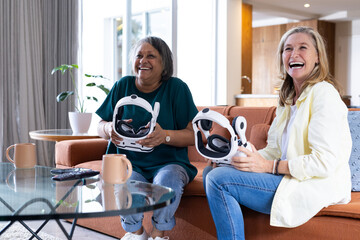 At home, holding vr headsets, two senior multiracial female friends enjoying virtual reality gaming