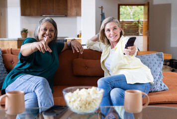 At home, watching tv and eating popcorn, senior multiracial female friends relaxing on couch