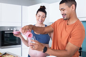 At home, Pouring smoothie into glass, pregnant multiracial couple smiling and enjoying healthy drink