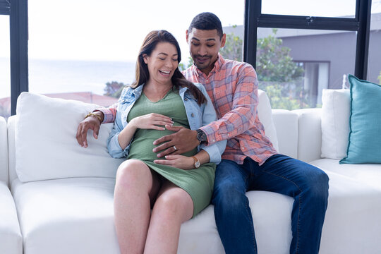 At home, Expecting multiracial couple sitting on couch, smiling and touching pregnant belly